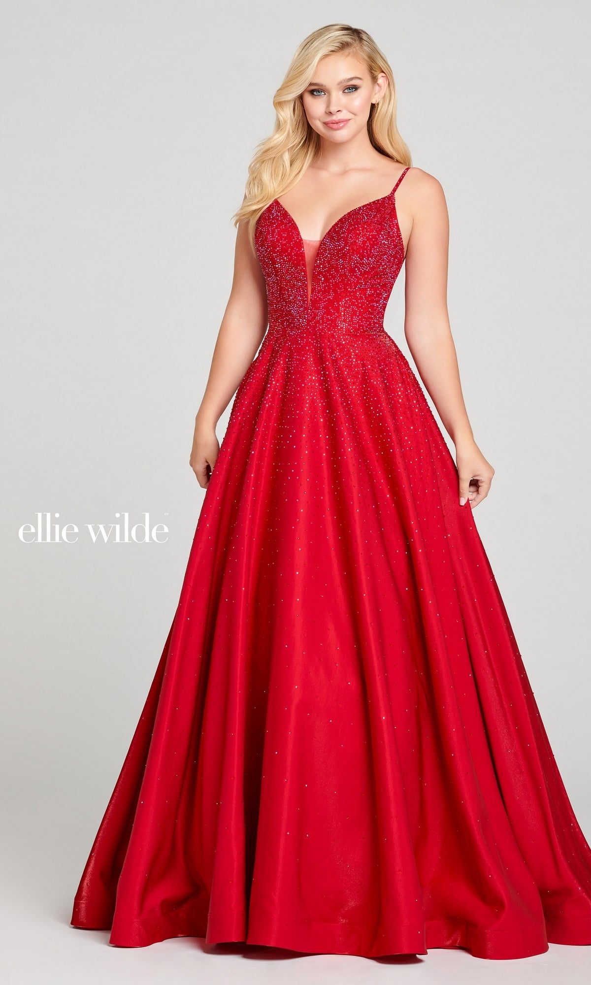 A woman stands against a plain background wearing the Ellie Wilde Ew121005, a sleeveless, floor-length red beaded prom ball gown with a deep V-neckline and subtle sparkle for an elegant finish.